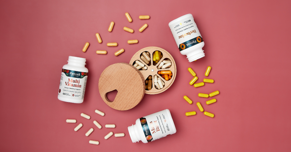 Flat lay image of three supplement bottles labeled "Berberine", "Multi Vitamin", and "Ma Rot" with loose capsules and a round wooden pill box on a pink background.