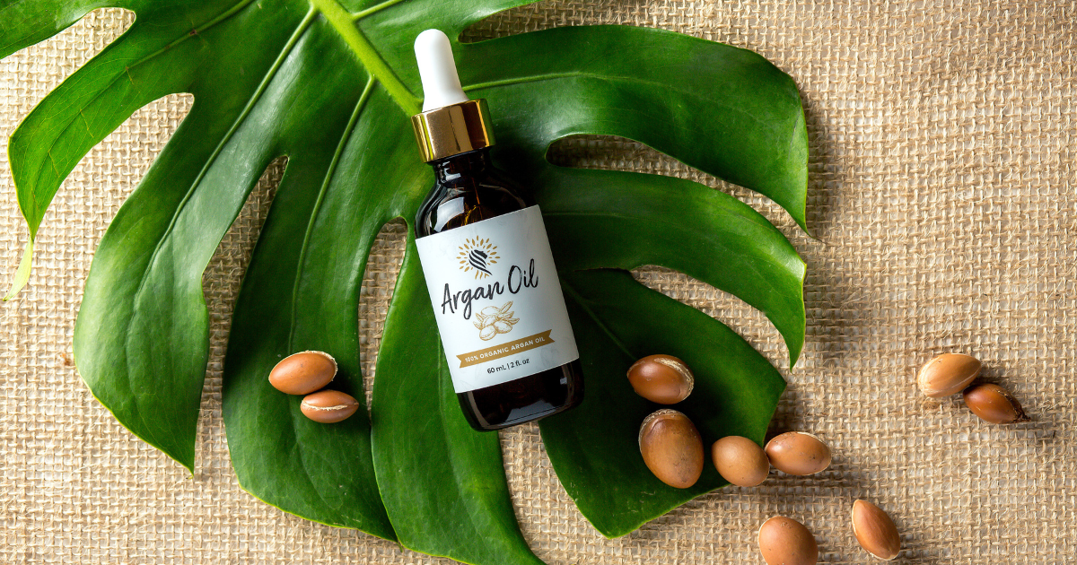 A dark glass bottle of "Argan Oil" with a dropper, surrounded by Argan nuts and a large green leaf on a burlap background.