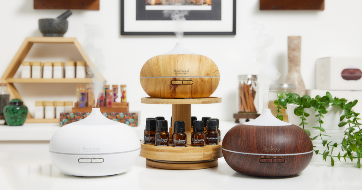 Three Beachwood Essentials diffusers, including a wooden diffuser on a stand, a white diffuser, and a dark wood diffuser, arranged on a table with essential oil bottles.