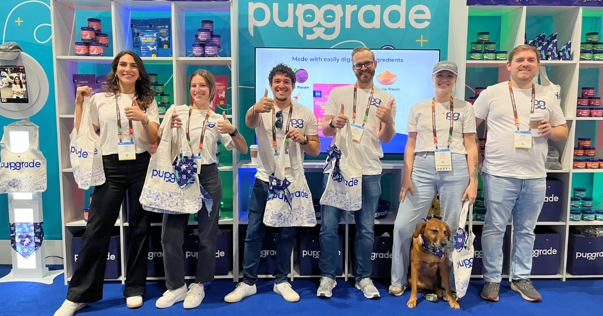 A group of six people, who appear to be of various ethnicities based on their skin tones, stand in front of a pet supply booth with the branding "pupgrade." They are all wearing white t-shirts with the "pupgrade" logo and are holding white tote bags with the same logo. A small dog, possibly a mixed breed based on its appearance, sits on the ground in front of them.