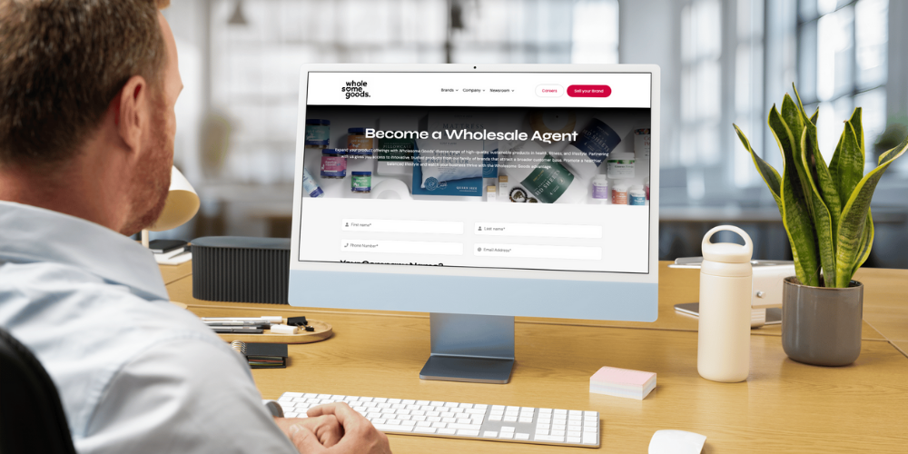 A person with short hair, viewed from behind, sits at a desk looking at a computer screen displaying a website with the text "Become a Wholesale Agent."
