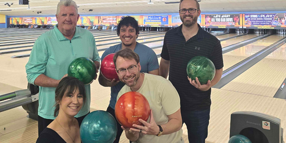 Six individuals of varying ages holding different colored bowling balls in a brightly lit bowling alley with multiple lanes visible in the background.