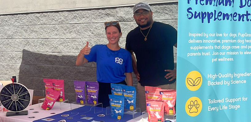 pupgrade walmart adoption event Two PupGrade team members smiling at an outdoor event booth with branded pet supplements, a prize wheel, and a promotional banner at Walmart’s Miami Pet Adoption Event.