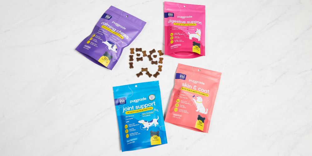 Four bags of Pupgrade dog supplements arranged on a white marble surface, surrounding a pile of bone-shaped treats. The bags are labeled "calming chews" (purple), "digestive support" (pink), "joint support" (blue), and "skin & coat" (coral).