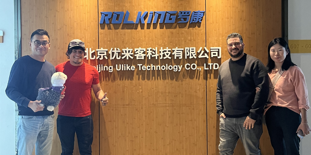 Four people from Wholesome Goods and a supplier stand smiling in front of the Beijing Ulike Technology Co., LTD office, with one person holding a plush alpaca mascot. The group poses in front of a wooden wall with the ROLKING logo and Chinese and English signage.