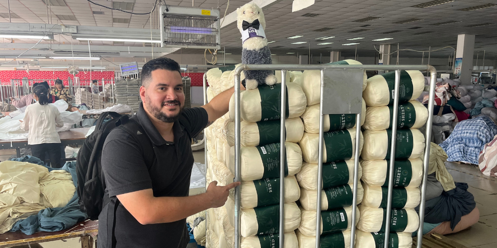 A Wholesome Goods team member poses inside a textile manufacturing facility next to a cart stacked with rolled Cosy House Collection bamboo sheet sets. A plush alpaca mascot sits on top of the stack, with factory workers and sewing stations visible in the background.