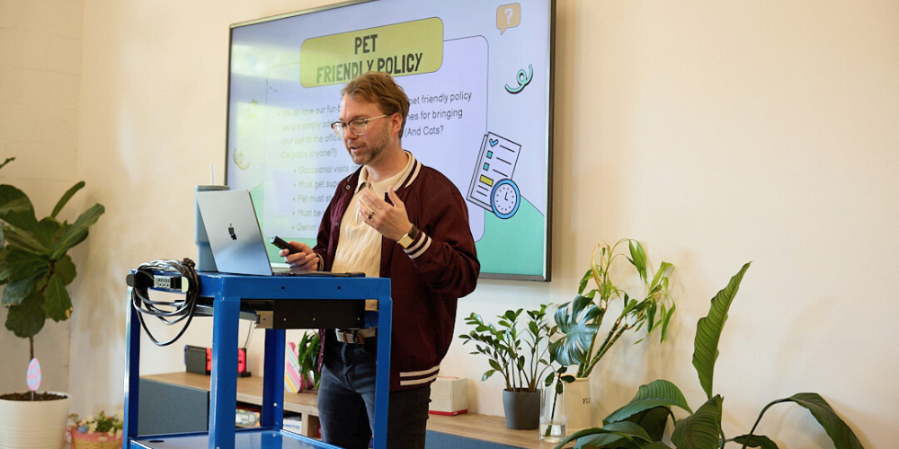 A Wholesome Goods team member gives a presentation on the company’s pet-friendly policy, standing beside a laptop cart and large screen, surrounded by greenery in a welcoming office space.