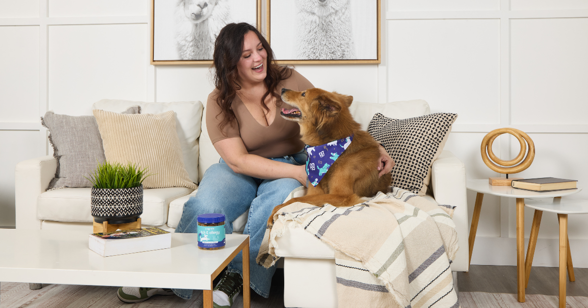 Smiling woman sitting on a cozy sofa at home with her happy dog wearing a blue bandana. A jar of PupGrade Itch & Allergy supplements sits on the coffee table. The scene captures a warm, relaxed work-from-home moment with pets.