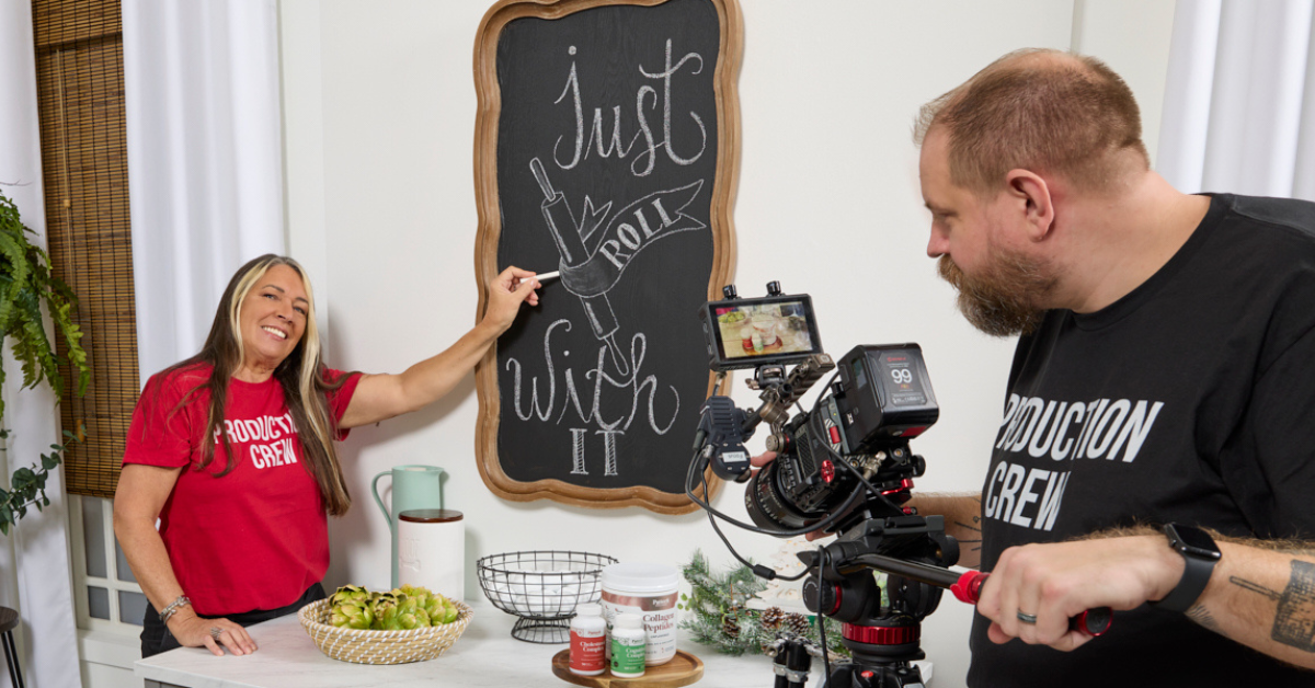 A member of the Wholesome Goods production team points to a chalkboard sign reading “Just Roll With It” while another team member films the scene with a professional camera in the studio.