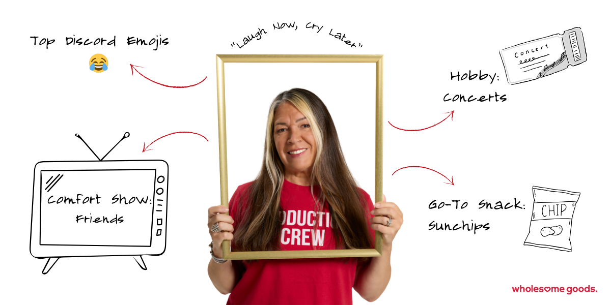 Senior Set Designer Lisa smiling while holding a picture frame, wearing a red “Production Crew” shirt at Wholesome Goods.